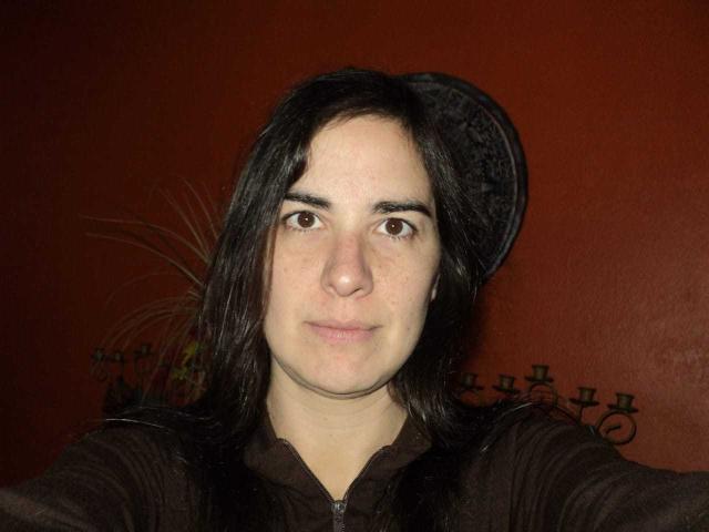 EstrellaHott with long black hair, brown eyes, and a neutral expression, wearing a dark brown zip-up, against a red wall, with a black hat slightly tilted.