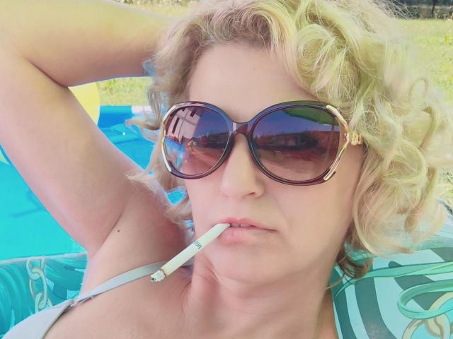 SexyCoco with curly blonde hair, wearing large brown sunglasses, has a cigarette in her mouth, and is lying down with an arm behind her head.