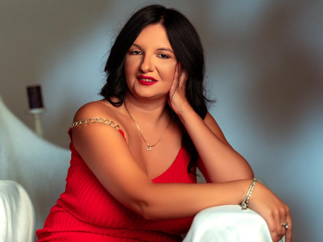 MadameAlexaX with wavy black hair, red dress, and gold chain necklace, sitting, hand on cheek, smiling, red lipstick, arm on white chair, bracelet, soft light.