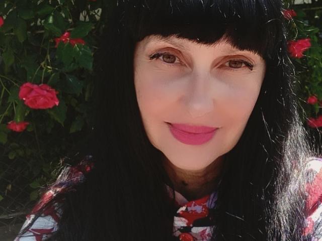 MelanyBanks has pale skin, brown eyes, black hair with bangs, and pink lipstick. She's wearing a red floral top and smiling at the camera. Her tits are average-sized.