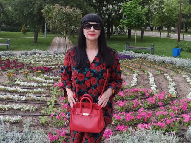 MelanyBanks with long black hair, wearing black sunglasses, red floral dress, and red purse, standing in front of colorful flower beds. She's got red lipstick and a cute ass.