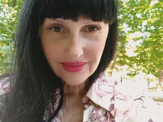 MelanyBanks has black hair, brown eyes, and pink lipstick. She's wearing a floral shirt, and her tits are partially visible. She's smiling, looking cute.