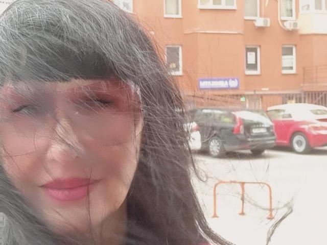 MelanyBanks with messy black hair, red lipstick, and slightly blurred face, standing in front of parked cars, wearing a dark shirt. Her tits are not visible.