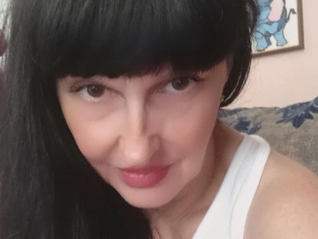 MelanyBanks has black hair with bangs, brown eyes, pink lipstick, and is wearing a white top. Her face is centered with a subtle smile. She has a cute, sexy look.