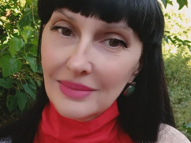 MelanyBanks with black hair, pink lipstick, green earrings, red turtleneck, and a slight smile, showing her nose and eyes. Her tits are not visible.