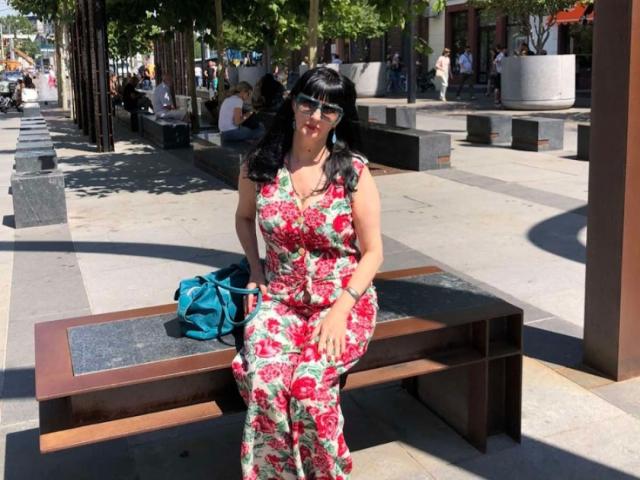MelanyBanks with black hair and bangs, wearing a floral dress, sitting on a bench, blue bag beside her, smiling, sunlit, urban setting, wearing sunglasses.