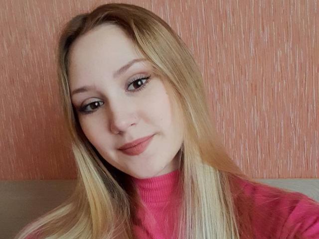 LaylaHott has blonde hair, light skin, and wears a pink turtleneck. She's smiling with a slight nose ring and subtle makeup, showing her tits in the photo.