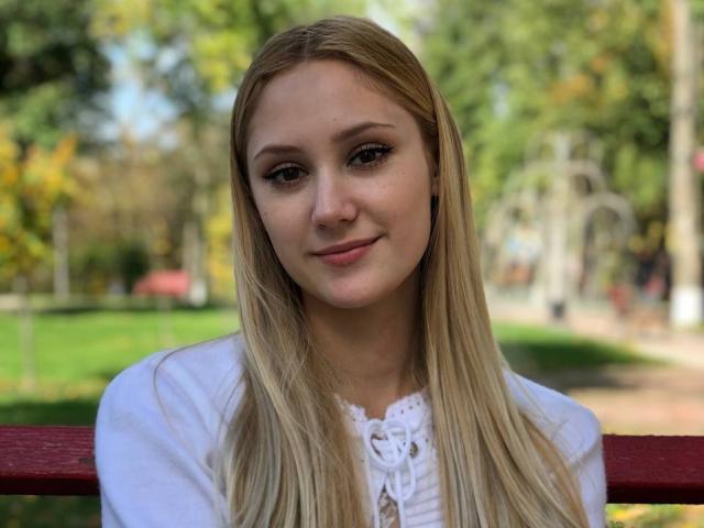 LaylaHott with long blonde hair, wearing a white shirt, smiling softly, standing in front of a red wooden railing, blurred green park background.
