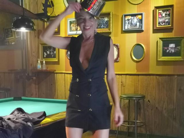 LunaBriella with small tits, wearing a black sleeveless dress with deep cleavage, holding a beer can over her head, smiling in a dimly lit bar, standing by a pool table.