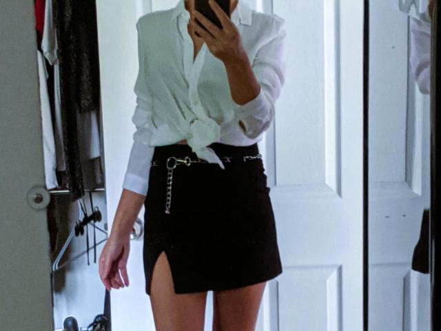 LunaBriella with a tied white shirt, black mini-skirt with a slit, and chain belt, holding a phone in front of a mirror, showing her bare legs and ass.