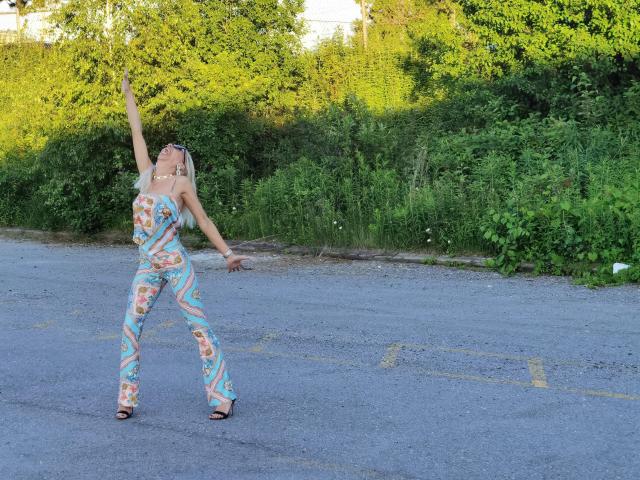 LunaBriella with blonde hair, wearing a colorful, floral, tight, one-piece outfit, and black sandals, dancing with arms raised on a gray asphalt road, surrounded by greenery.