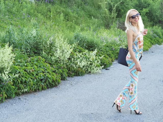 LunaBriella with blonde hair, wearing a colorful, patterned, sleeveless jumpsuit, black heels, and sunglasses, walking on a gravel path, holding a black clutch.