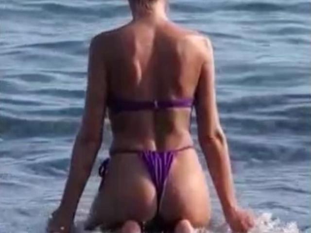 LunaBriella is kneeling in water, facing away, wearing a purple thong bikini that shows her ass and small tits. Her back is tan and she's in sunlight.