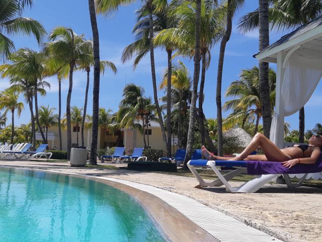 LunaBriella, a hot chick with a tan, is lounging on a blue chaise by the pool, wearing a black bikini, with her ass and tits exposed, under palm trees.