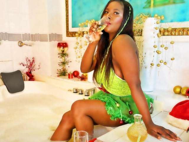 MollyGoldenn with dark skin, long green braids, green lace top, green skirt, sitting in a white bathtub, drinking champagne, showing her tits, with a glass and bottle on the tub.