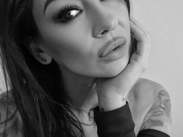 Black-and-white photo of Xentthia with long dark hair, pouty lips, tattoo on right arm, hand near face, wearing a zipped-up shirt. Her eye is slightly up.