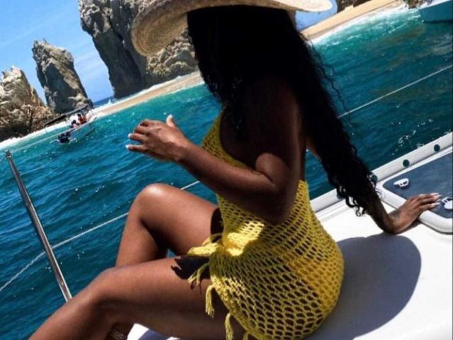 KatyBomb with dark skin, long curly black hair, wearing a yellow fishnet dress and straw hat, sitting on a boat, showing her ass and legs, with blue water.