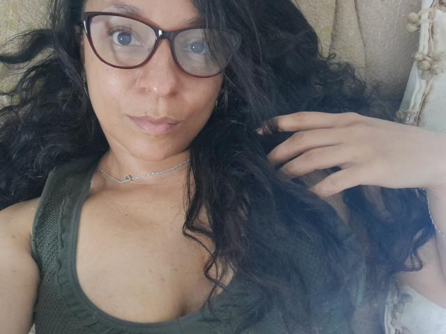 Angelonix has curly black hair, wearing glasses, a green tank top, and a silver necklace, lying down with a hand in her hair, looking at the camera.
