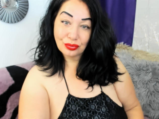 LexyRose has red lipstick, black hair, and thick eyebrows. She's wearing a black halter top with a pattern, showing her bare shoulders and a little of her tits.