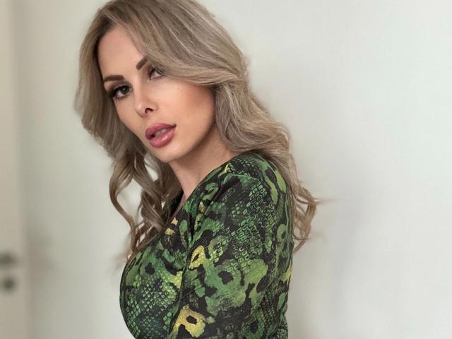 XANIA-WET tem cabelos loiros ondulados, uma blusa verde com estampa de cobra e lábios rosados e carnudos. Seus seios são de tamanho médio e ela está olhando por cima do ombro com uma expressão sedutora..