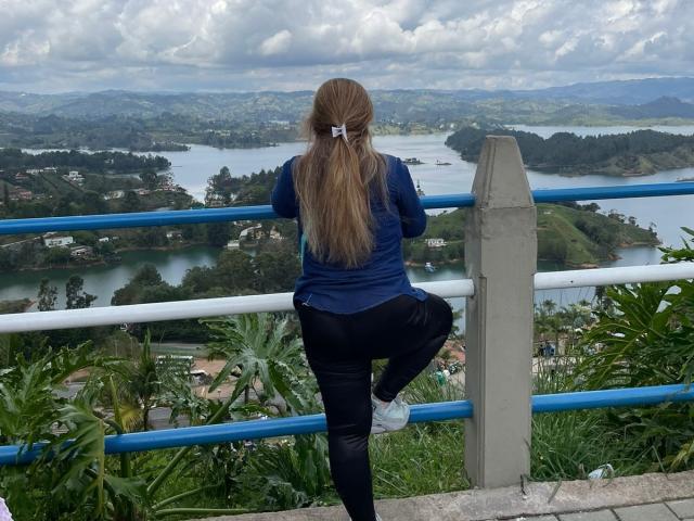 GilliSkquirtty with long blonde hair in a blue shirt and black jeans, leaning on a blue and white railing, showing her ass, looking over a scenic, green landscape.