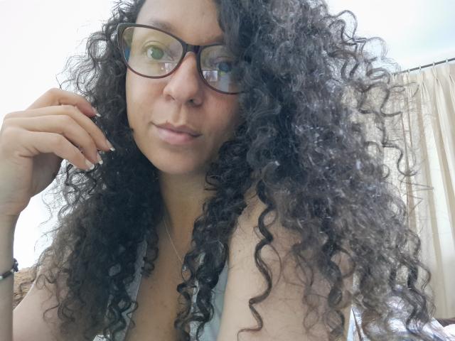 Angelonix with curly black hair, glasses, and a slight smile, showing her small tits in a low-cut top, resting her hand on her cheek.