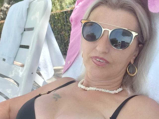 SexyCoco with blonde hair, gold sunglasses, black bra, white shell necklace, gold hoop earrings, small tattoo on right breast, pink towel behind, white chair, and relaxed smile.