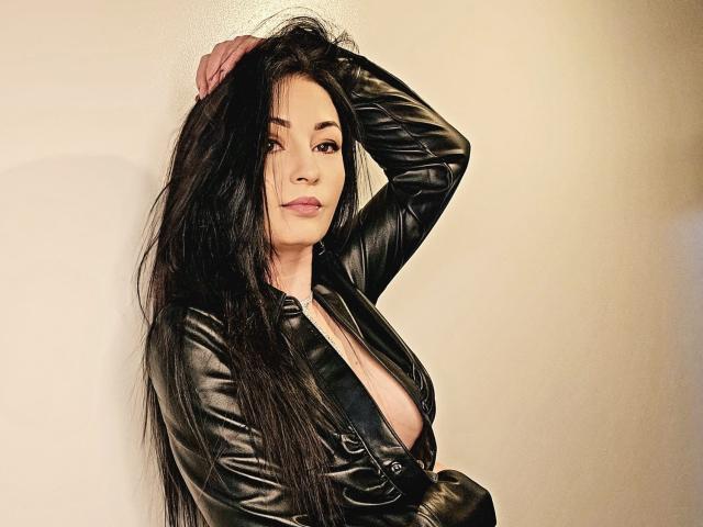 Coryna with long black hair, wearing a black leather jacket, exposing her tits, hand in hair, looking at camera, soft light, slightly parted lips, confident expression.