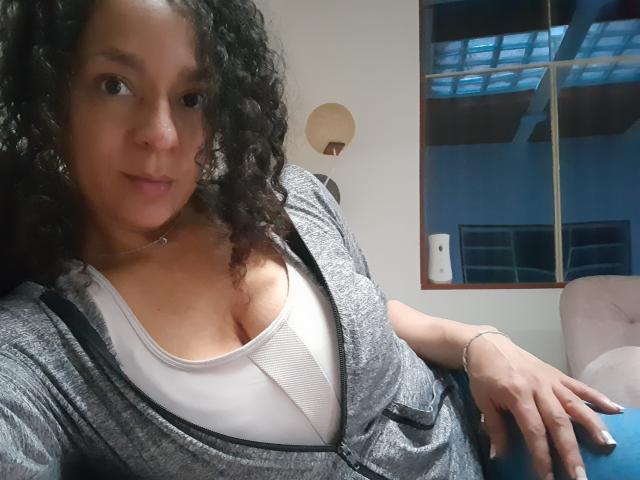 Angelonix with curly black hair, light brown skin, and medium-sized tits in a grey zip-up and white top, sitting, smiling, wearing a necklace, and a bracelet, in a relaxed setting.