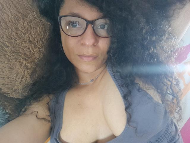 Angelonix with curly black hair, wearing glasses, grey tank top, showing cleavage, lying down, brown fuzzy pillow, necklace, looking at camera, medium skin, subtle smile.