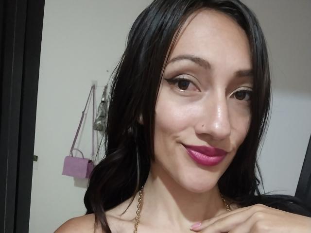 KiriamGomez has dark hair, pink lipstick, and a small beauty mark on her right cheek, smiling with brown eyes and a gold necklace. Boobs not visible.