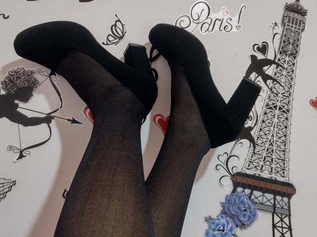KiriamGomez's legs in black sheer stockings and black high-heeled shoes with ankle straps, feet crossed, against a Paris-themed wall.