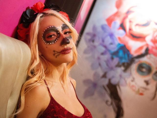 Blonde KiriamGomez with face paint, red lace top, black bow, and red hair, sitting on beige chair, looking at colorful wall art. Boobs, painted face, sexy, cute.
