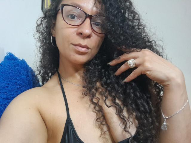 Angelonix with curly black hair, glasses, and a black halter top, showing a ring on her right hand and a silver bracelet, looking at the camera. Boobs visible.