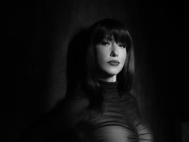 Black and white photo of MissVonTease with dark, straight hair, bangs, and serious expression, wearing a ruffled, sheer top, highlighting her tits subtly. Dark background.