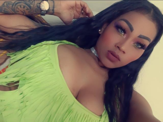 LinaaPrincess has green eyes, thick eyebrows, and long black hair. She's wearing a neon green dress with fringes, showing her big boobs. She's looking at the camera.
