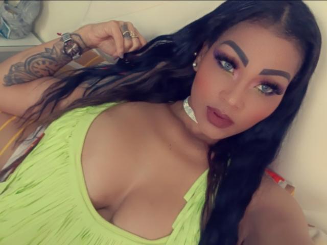 LinaaPrincess has big tits in a neon green dress, long black hair, blue eyes, bold eyebrows, red lipstick, white choker, tattoo on arm, and is looking at the camera.