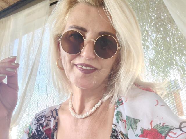SexyCoco with blonde hair, wearing round sunglasses, floral top, pearl necklace, and dark lipstick, smiling, holding curtain, showing her tits slightly, in bright daylight.