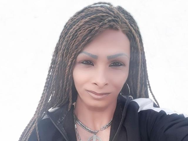 SiliconBoobs has brown braided hair, dark eyebrows, and is wearing a black jacket with white stripes. She's smiling with a silver necklace and hoop earrings.