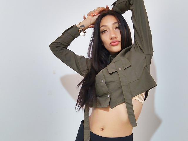 KiriamGomez with long black hair, green military-style jacket, and raised arms, exposing her small tits and flat stomach. She's wearing a beige bra and black pants.
