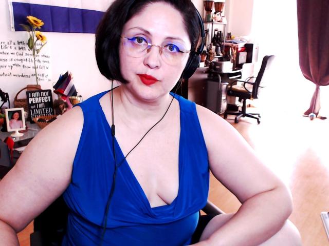 ImperatrizaSanda with pale skin, black bob, glasses, red lipstick, blue dress, showing big tits, sitting, headphones on, in a room with a desk and chair.