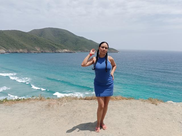 SamantaDark with braided hair, wearing a blue dress, standing on a rocky cliff, holding a small white object, looking at the camera, with a blue ocean and green hills.