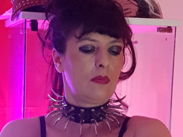 MaitresseElsa with dark hair, heavy makeup, red lipstick, black spiked choker, and black strap dress, looking down with a serious expression, pink and red lighting.
