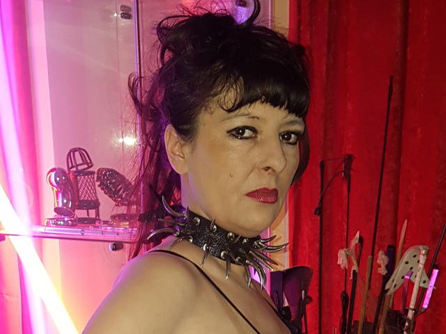 MaitresseElsa with dark hair, bangs, red lipstick, spiked collar, and bare shoulders, looking at camera, surrounded by neon pink and red lighting, with musical instruments in background.