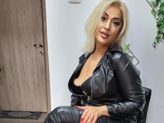 SexyMilf with blonde hair, full tits in a black leather zip-up, sitting, looking at camera, red nails, wooden door in background, plant on right, confident, sexy.