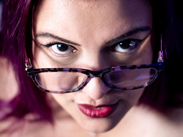 SammyOwens with purple hair, tortoiseshell glasses, and red lipstick, staring intensely with her big brown eyes. Her tits are partially visible, and she's looking right at the camera.