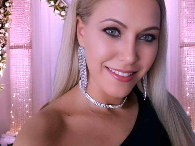 MyaHott has blonde hair, blue eyes, and is smiling at the camera. She's wearing large diamond earrings and a matching choker. She's dressed in a black, one-shoulder outfit.