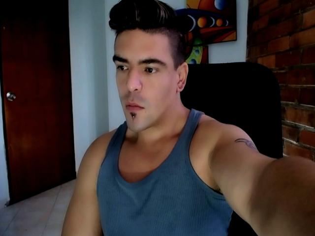 MatthewRay has a muscular build, wearing a blue tank top, short dark hair, a goatee, and a tattoo on his right arm, taking a selfie in a dimly lit room.
