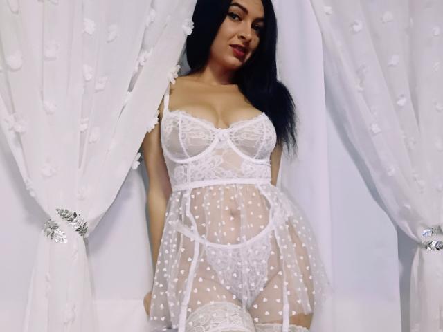 PassionX with long black hair, wearing sheer white lace lingerie, showing her tits and pussy, standing in front of white floral curtains, smiling slightly.