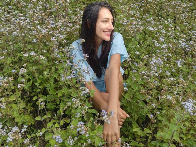 KiriamGomez with long black hair, blue shirt, squatting in green field, surrounded by small white flowers, smiling, barefoot, hands on knees, looking to the side.